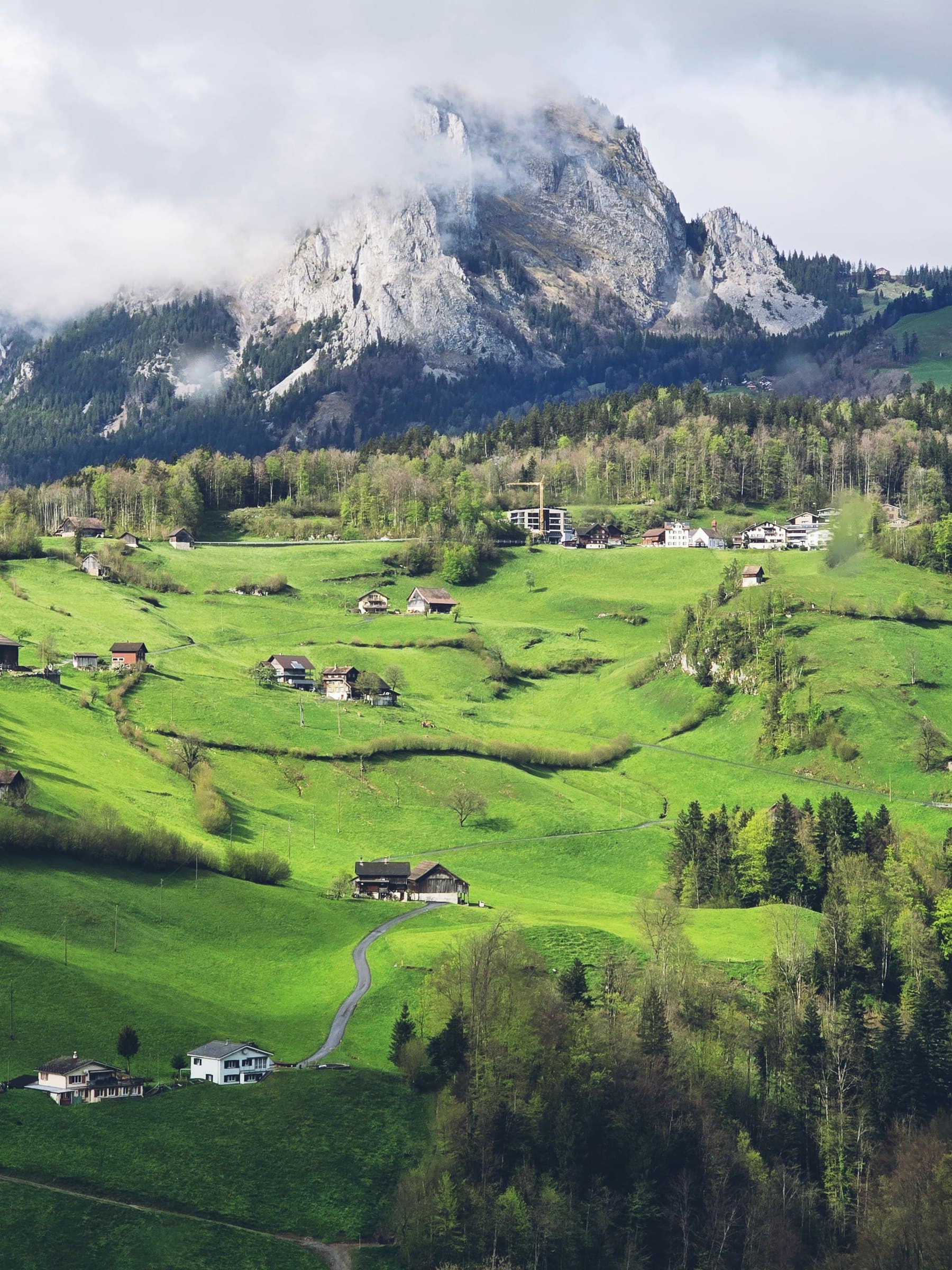 An alpine valley in Switzerland — scattered farmhouses on green pasture below a limestone peak in cloud.
